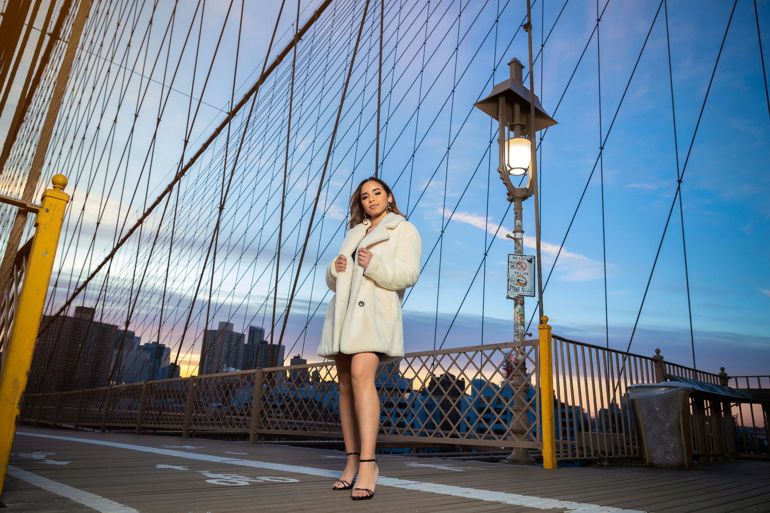 Woman in white coat poses on Brooklyn Bridge at dusk with city skyline and lamp post behind her.