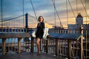 Woman in black dress poses on Brooklyn Bridge at sunset with Manhattan skyline in background.