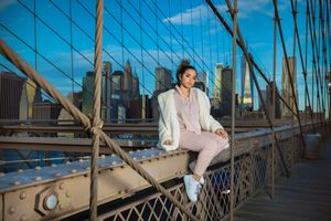 Woman in white jacket sits on Brooklyn Bridge railing with NYC skyline at sunset.