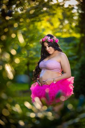 Pregnant woman in pink tutu skirt and floral crown cradling baby bump outdoors in lush green garden setting.