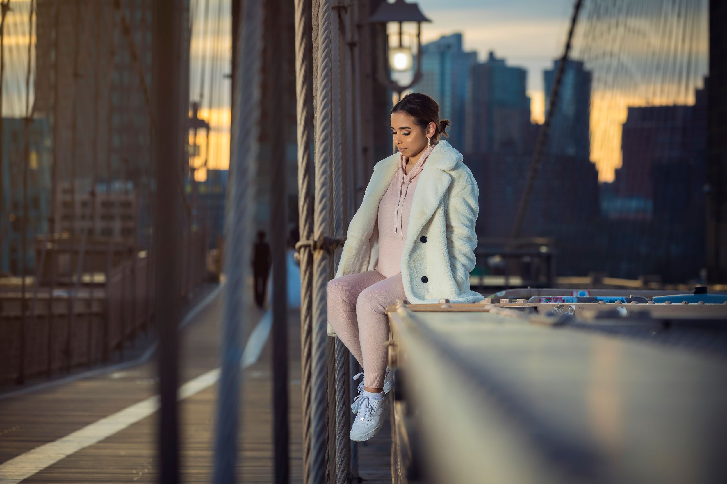 Woman in white coat sitting on Brooklyn Bridge railing at sunset with NYC skyline in background.