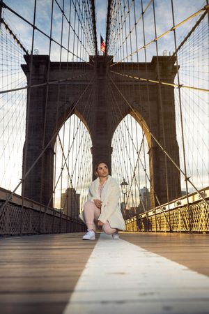 Woman posing on Brooklyn Bridge walkway at sunset, with iconic stone towers and suspension cables in background.