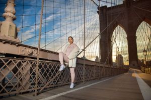 A woman poses gracefully on the Brooklyn Bridge walkway at sunset, wearing a pink outfit and white sneakers.