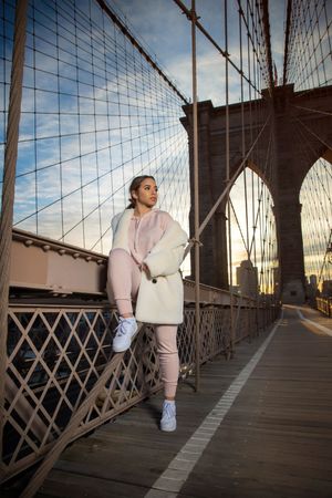 Woman in white coat and pink outfit posing on Brooklyn Bridge at sunset.