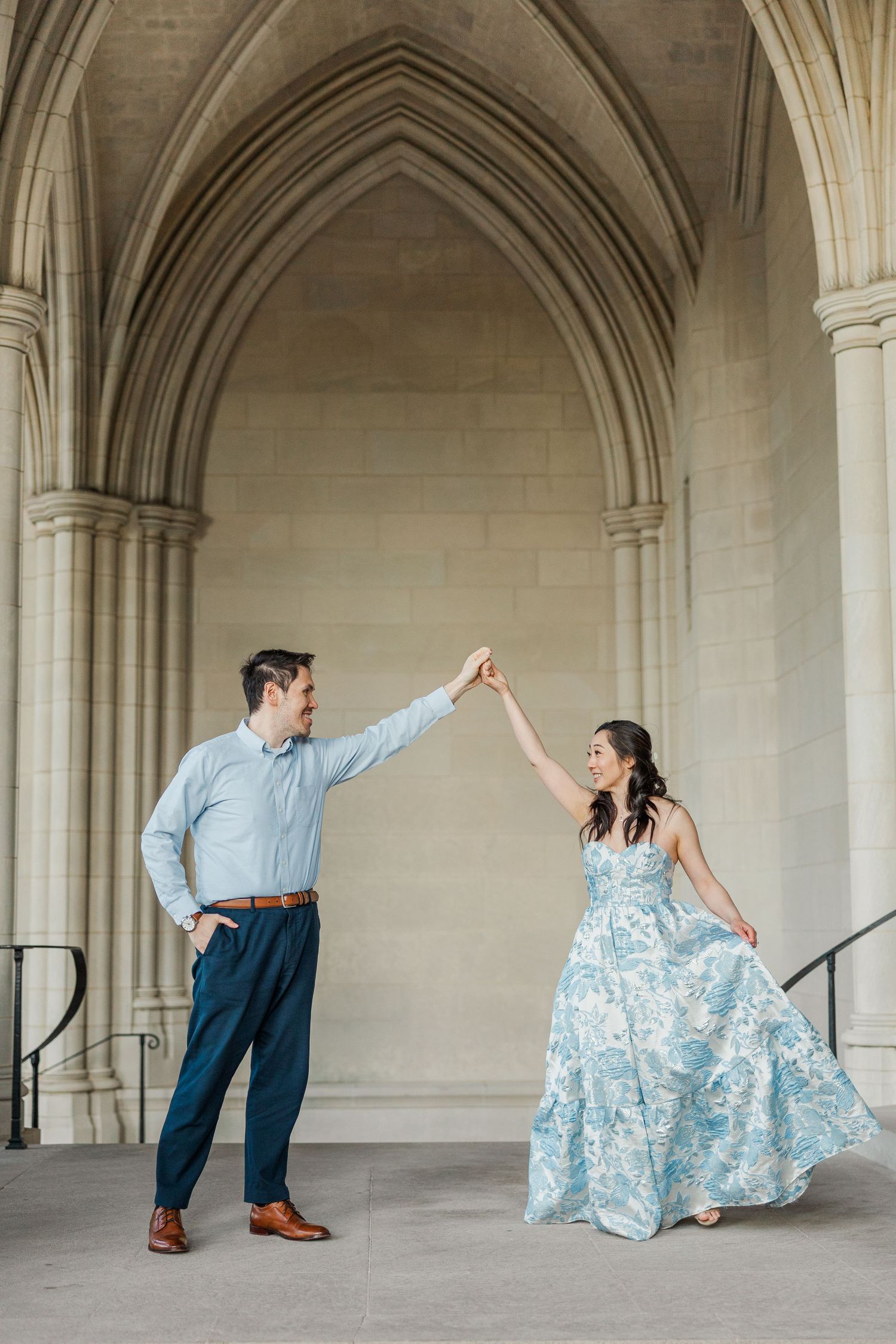 Couple dancing under Gothic stone arches, woman in blue floral gown, man in blue shirt and navy pants, elegant venue.