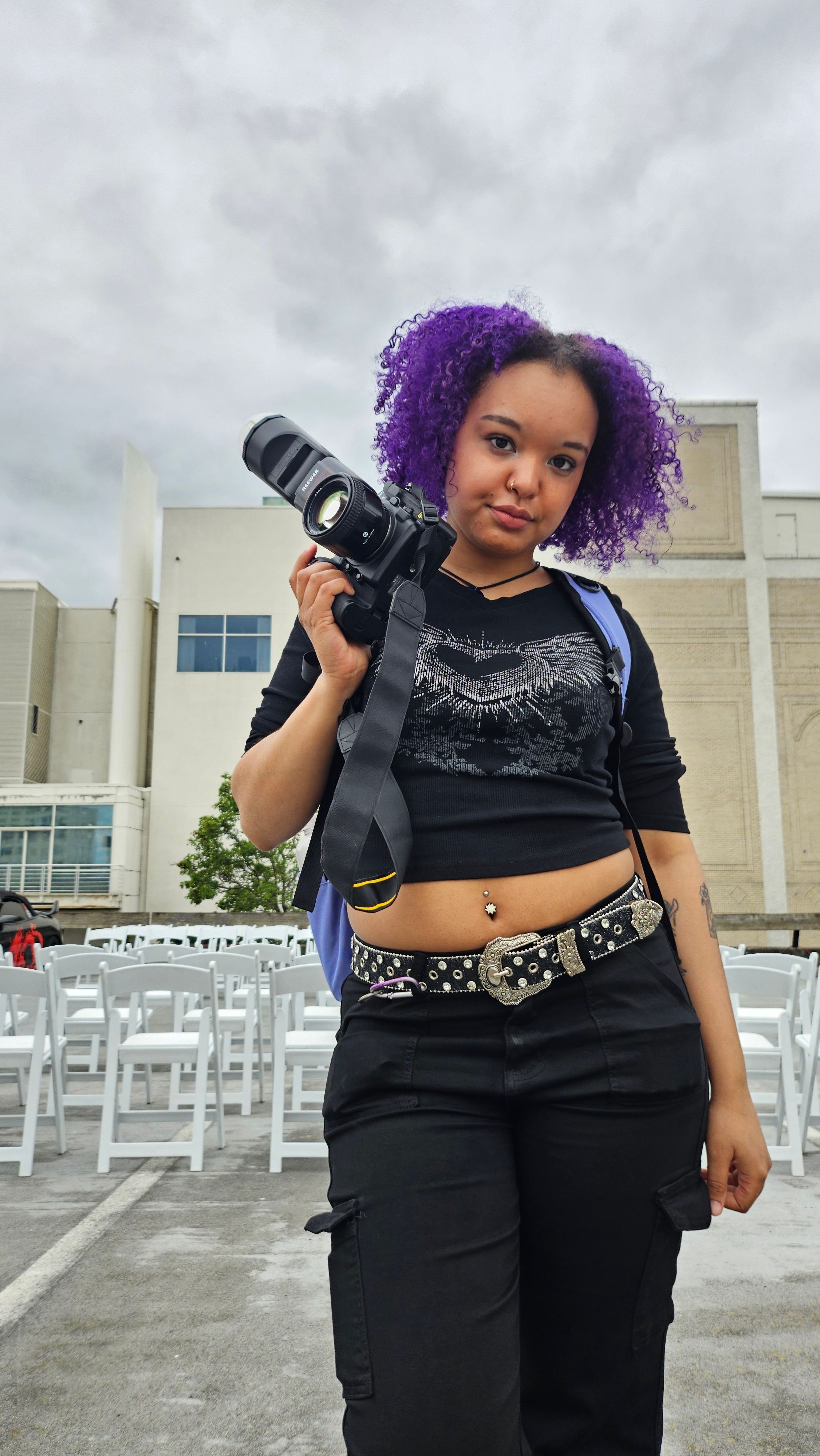 Young woman with purple curly hair holding a professional camera outdoors, wearing black crop top and cargo pants.