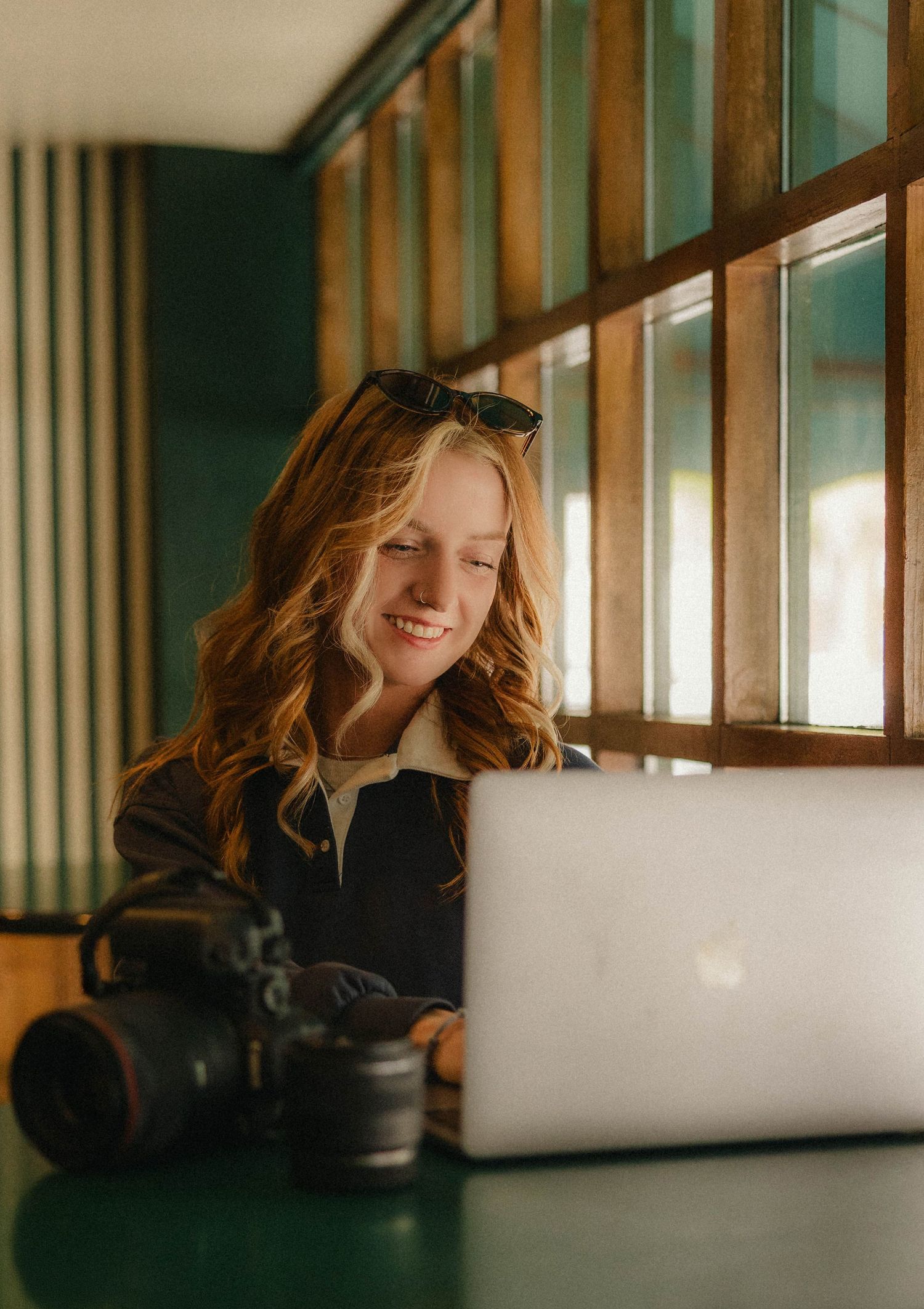 Smiling young woman with wavy blonde hair working on a MacBook laptop beside a DSLR camera in a cozy cafe.