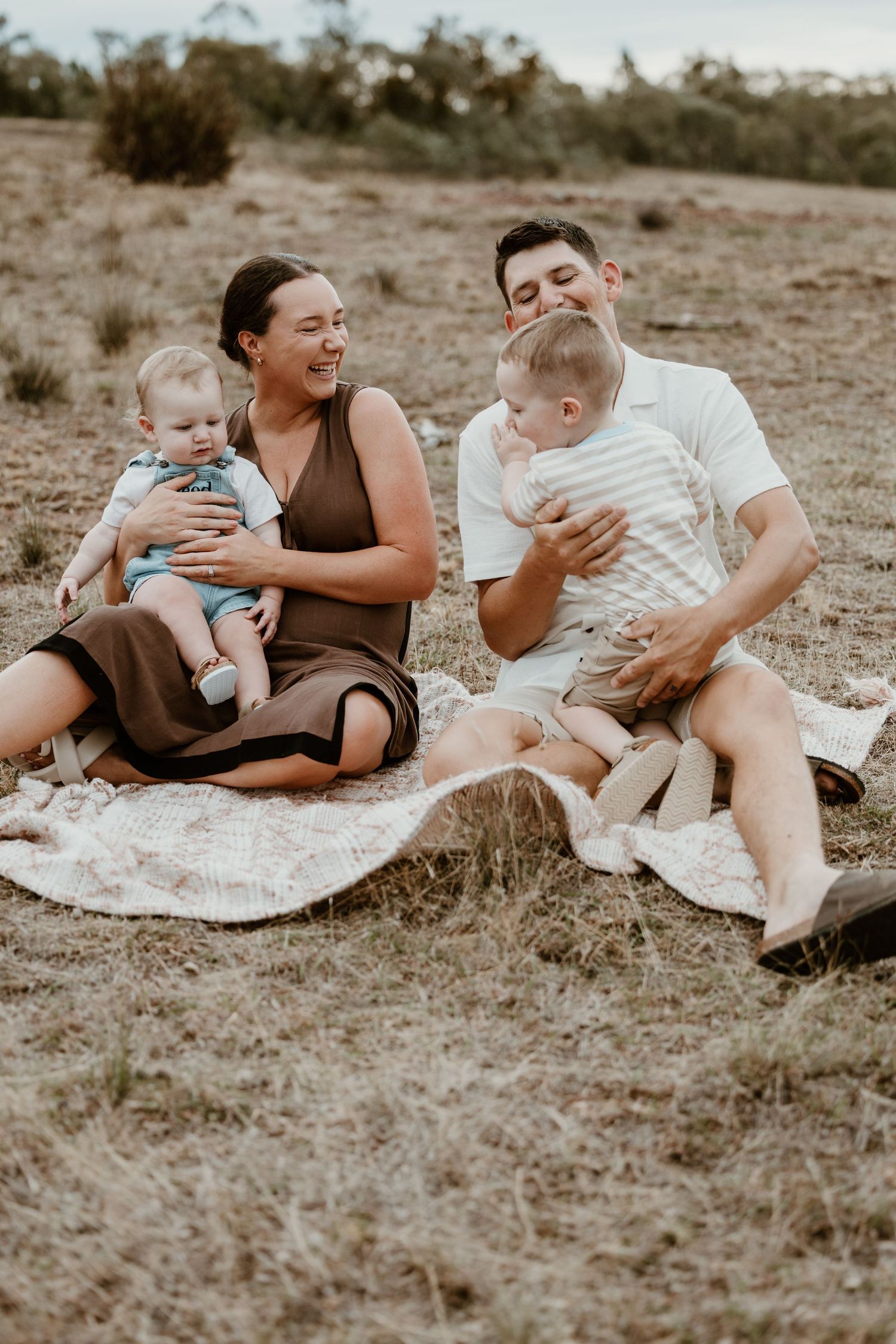 Family of four sitting on a blanket in a field, parents laughing with two young children during an outdoor picnic.