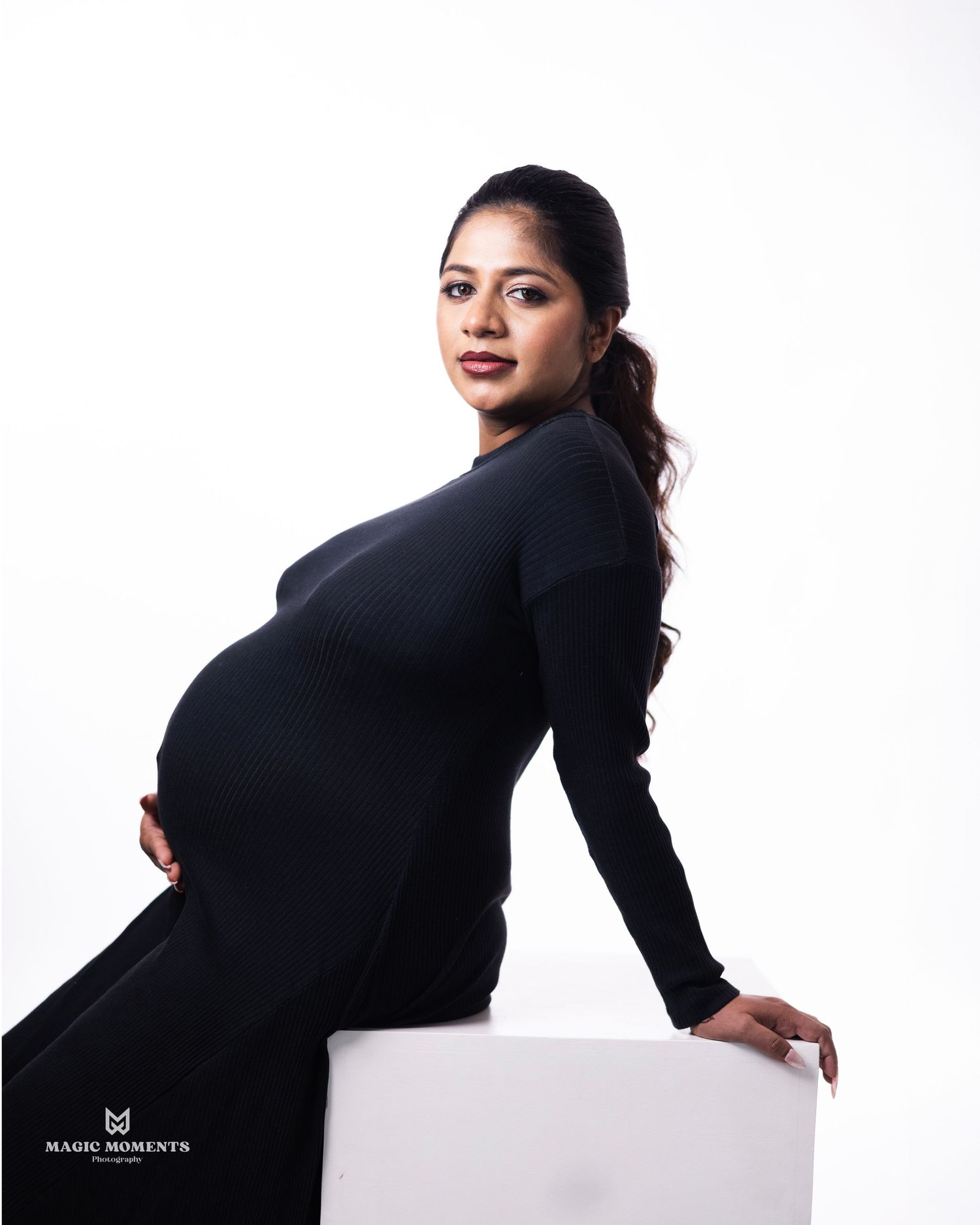 Pregnant woman in black dress posing for maternity photoshoot against white background.