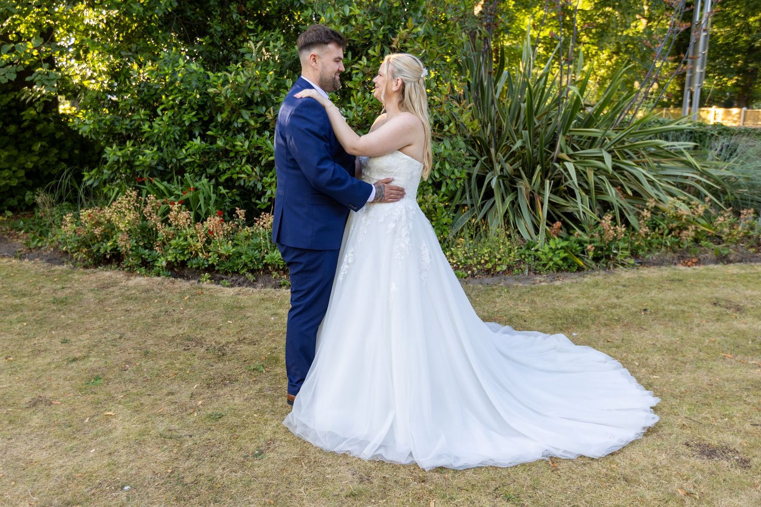 A bride in a white gown and groom in a navy suit share a romantic moment in a lush garden setting.