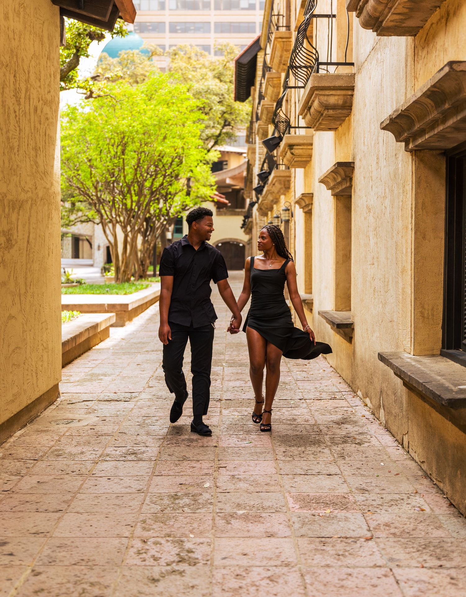 Couple in black outfits holding hands while walking along a charming cobblestone alley flanked by historic buildings.