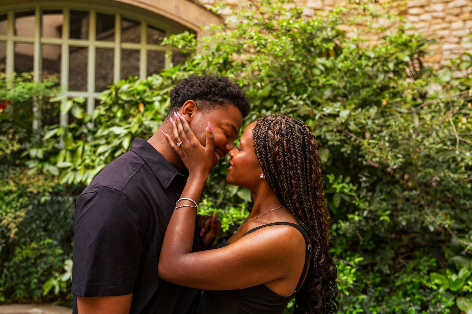 A young Black couple shares a romantic almost-kiss moment outdoors, surrounded by lush green foliage.