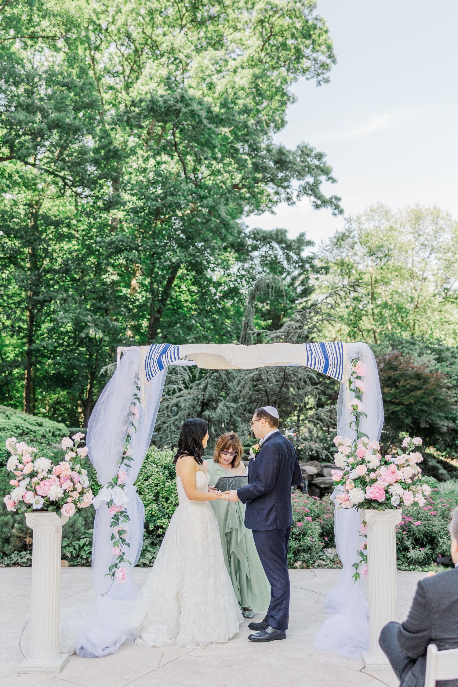Outdoor Jewish wedding ceremony under a floral chuppah with pink roses, bride and groom exchanging vows surrounded by lush greenery.