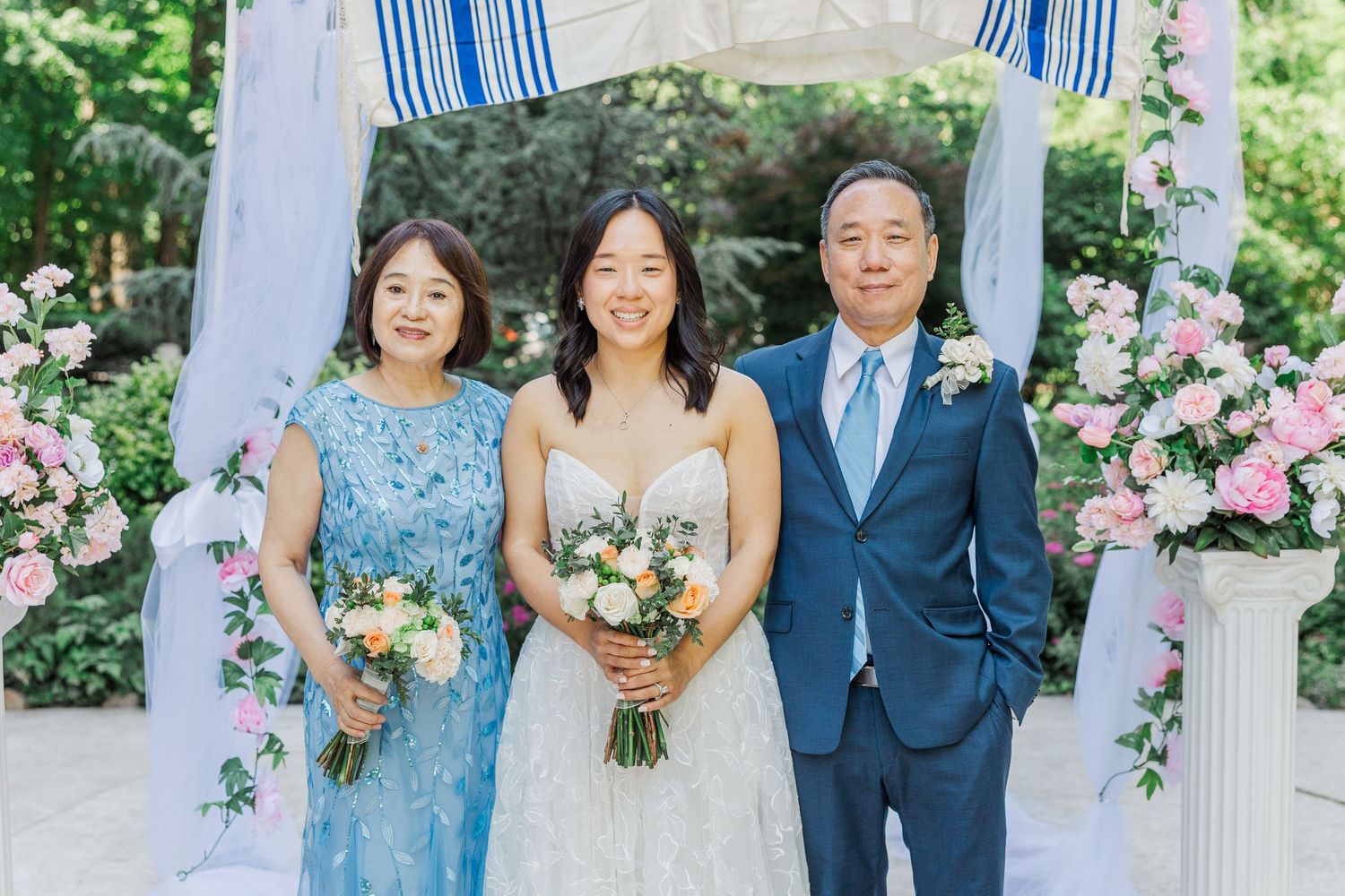 Bride in white gown poses with parents in blue attire at outdoor wedding ceremony with pink floral arrangements.