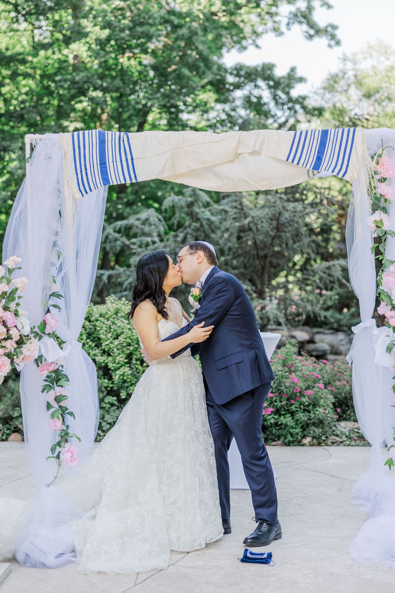 Bride and groom share first kiss under floral chuppah with blue striped tallit at outdoor Jewish wedding ceremony.