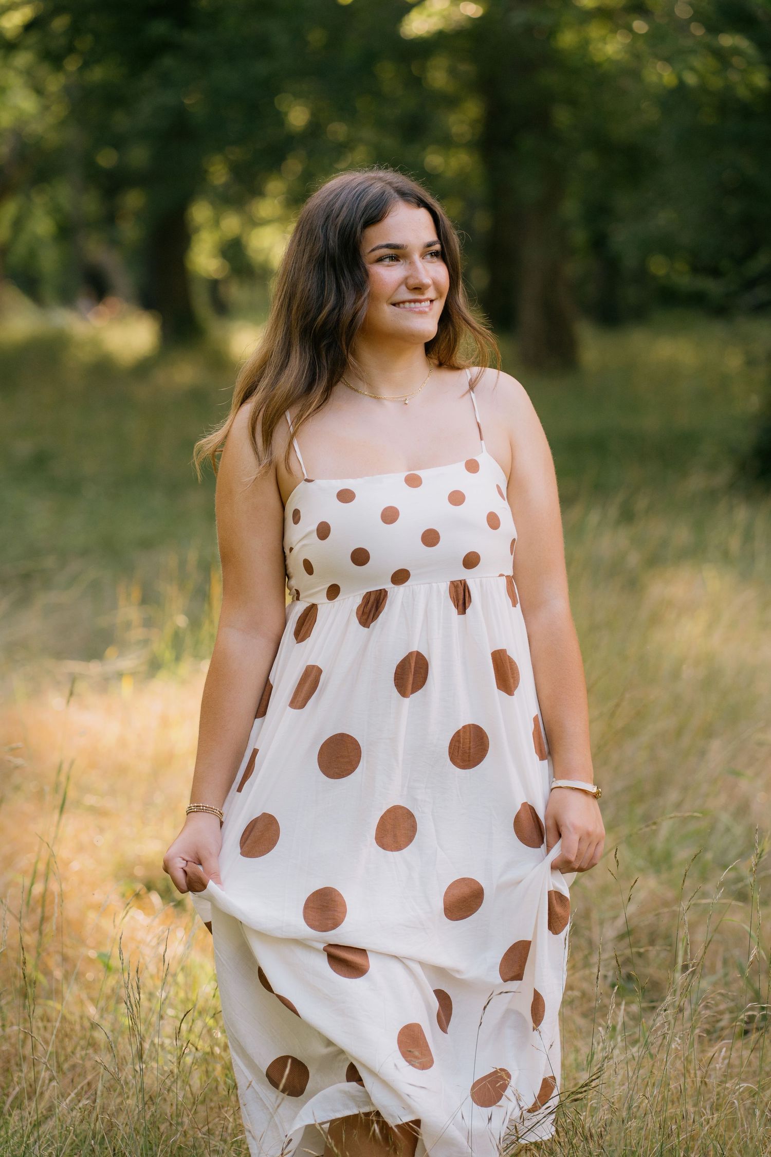 Young woman in white polka dot sundress smiling in a sunlit field with green trees in background.