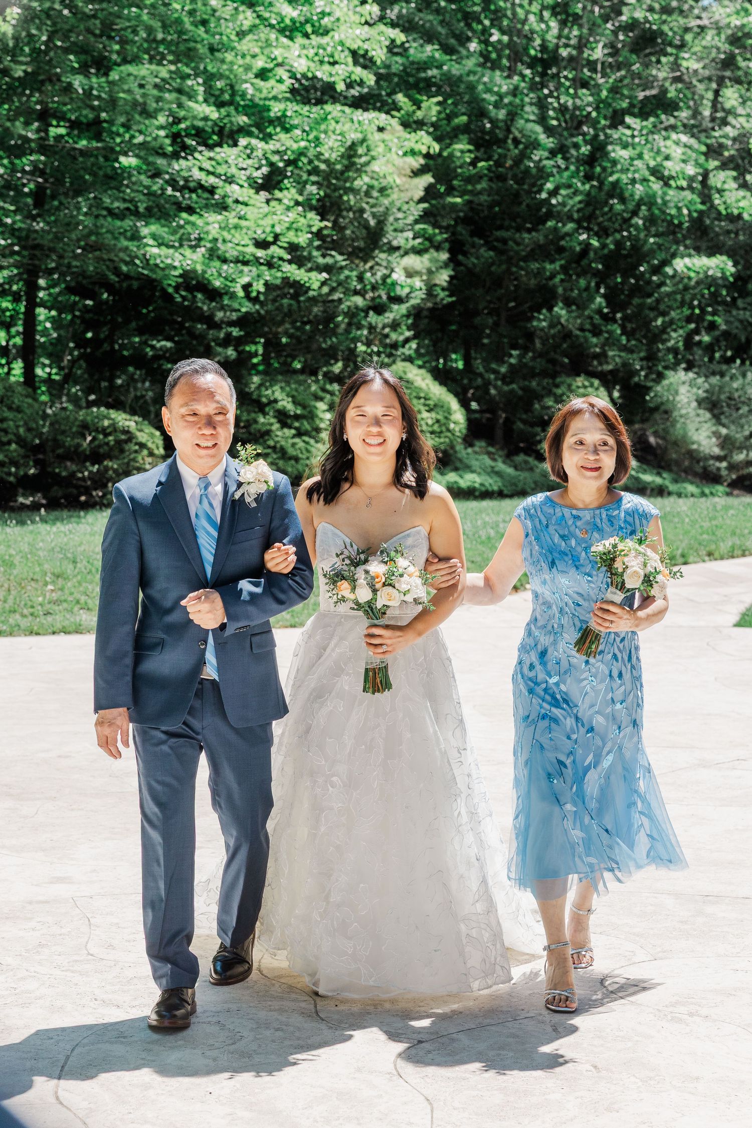 Bride in white gown walks with parents in blue attire, holding bouquets outdoors on sunny day.