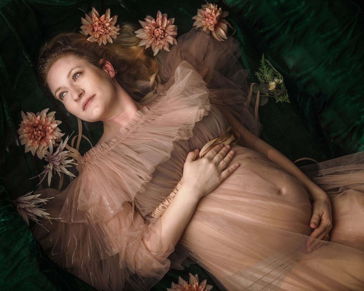 Pregnant woman in sheer blush gown lying among pink dahlias on green velvet, elegant maternity portrait.