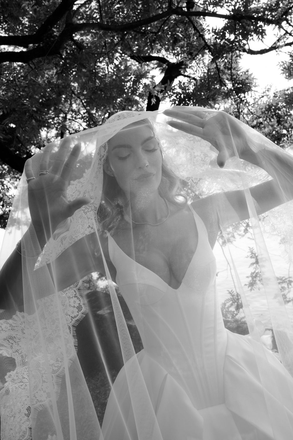 Black and white photo of a bride lifting her lace veil outdoors under a tree in an elegant wedding gown.