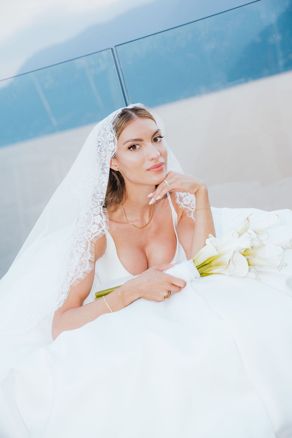 Elegant bride in white gown and lace veil holds calla lily bouquet, posing gracefully outdoors.