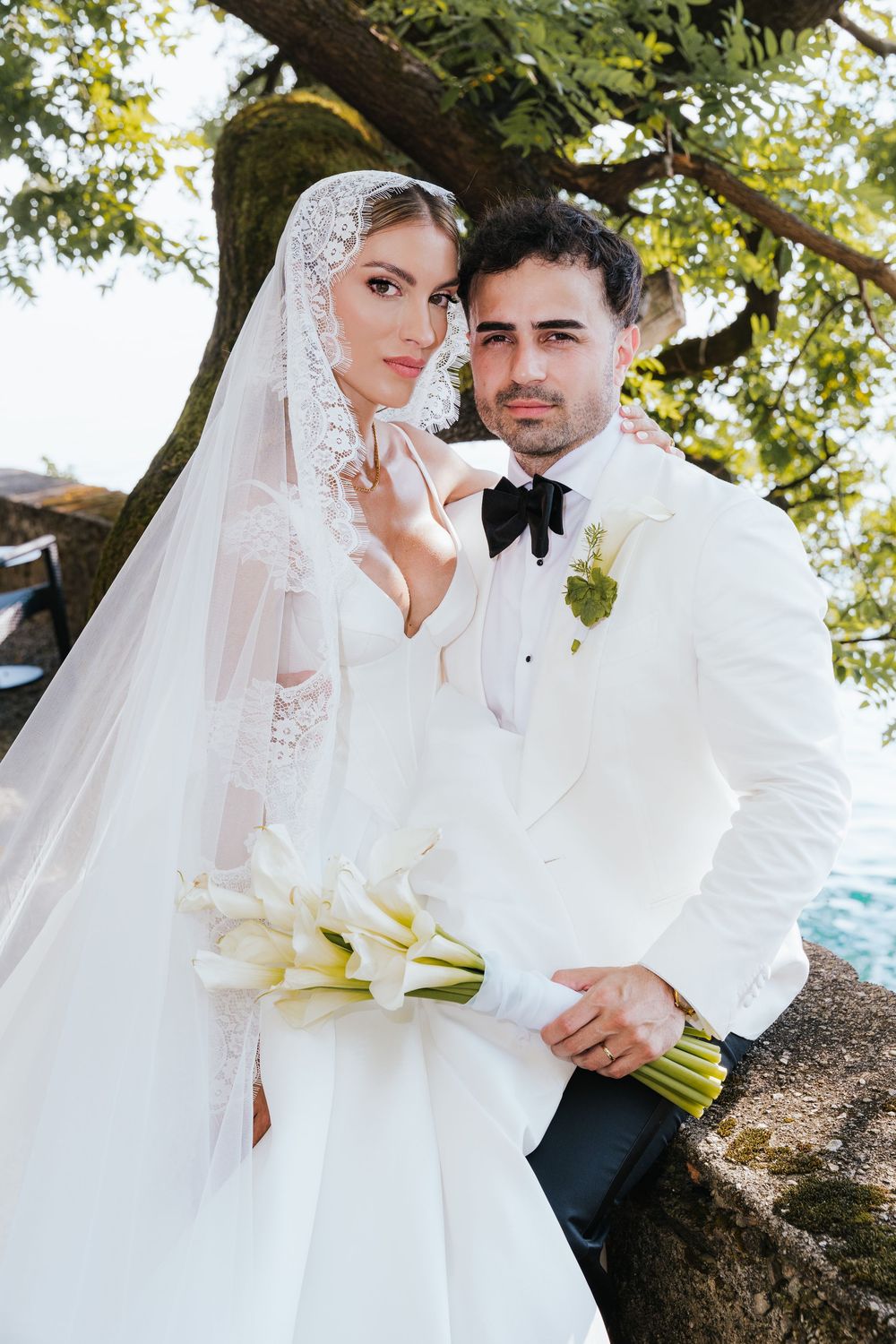 Elegant bride and groom pose under a tree, she wears a lace veil and holds white calla lilies, he wears a white tuxedo.