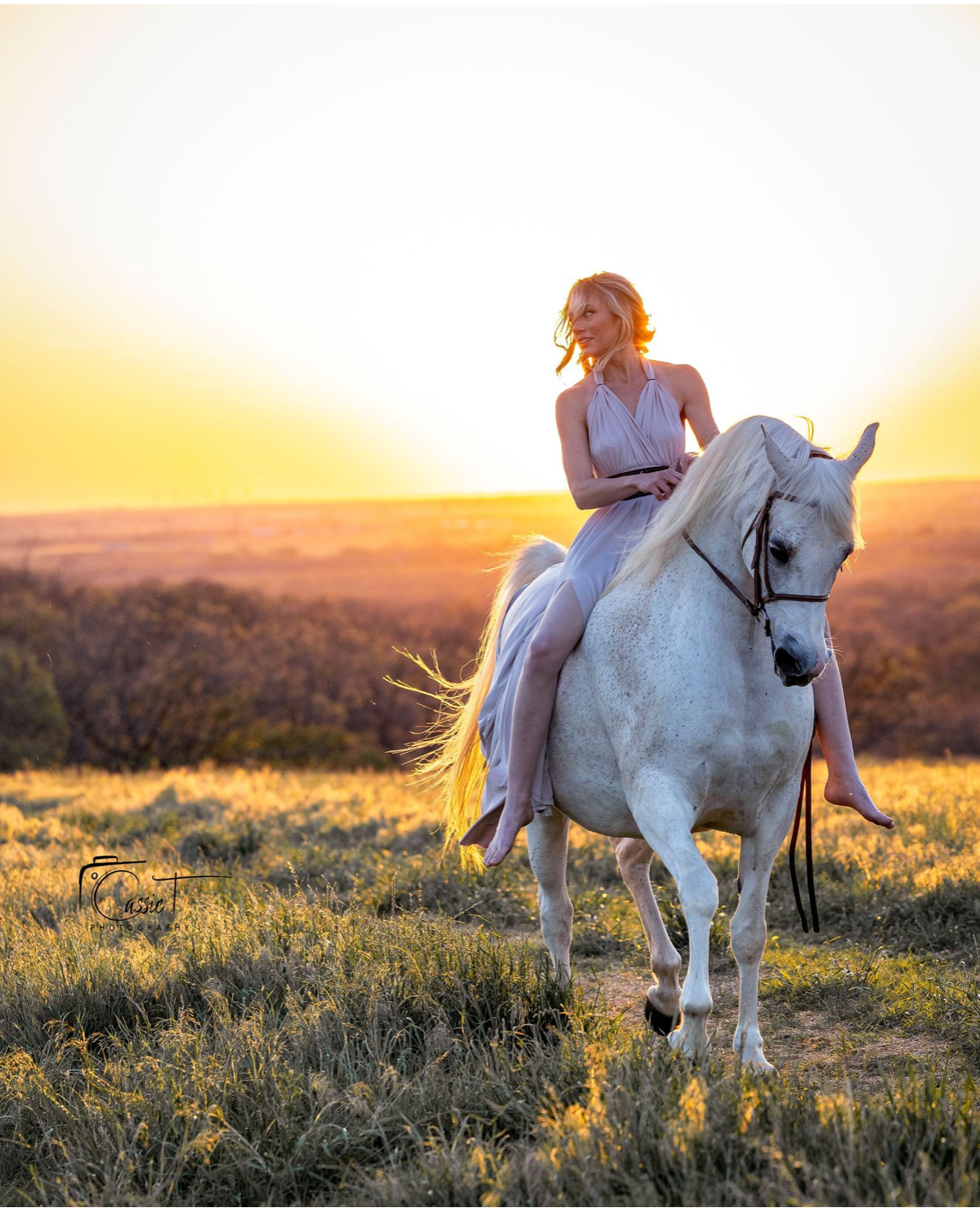 Woman in pink dress riding white horse at golden sunset over scenic landscape.