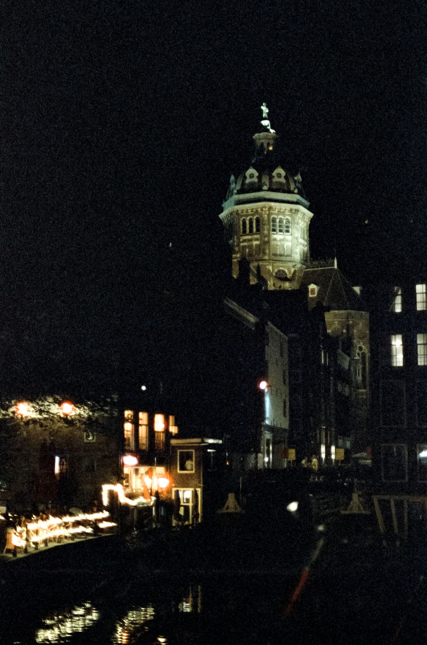 Illuminated historic tower and canal buildings reflected in water at night in Amsterdam, Netherlands.