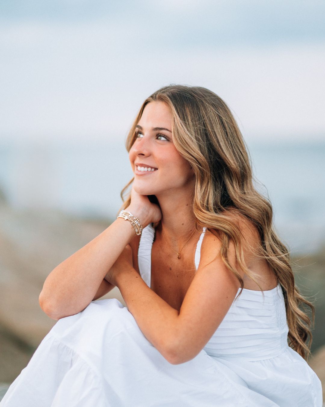 Young woman in white dress sitting by the ocean, smiling with wavy blonde hair and bracelets on a sunny day.