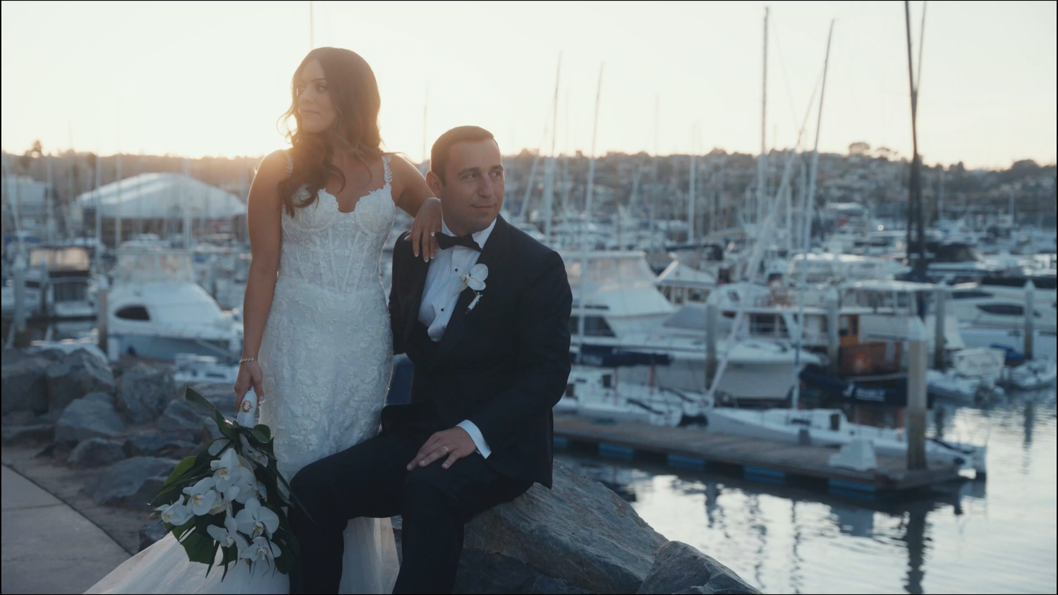 A bride and groom pose romantically at a marina during golden hour, with boats and a coastal skyline in the background.