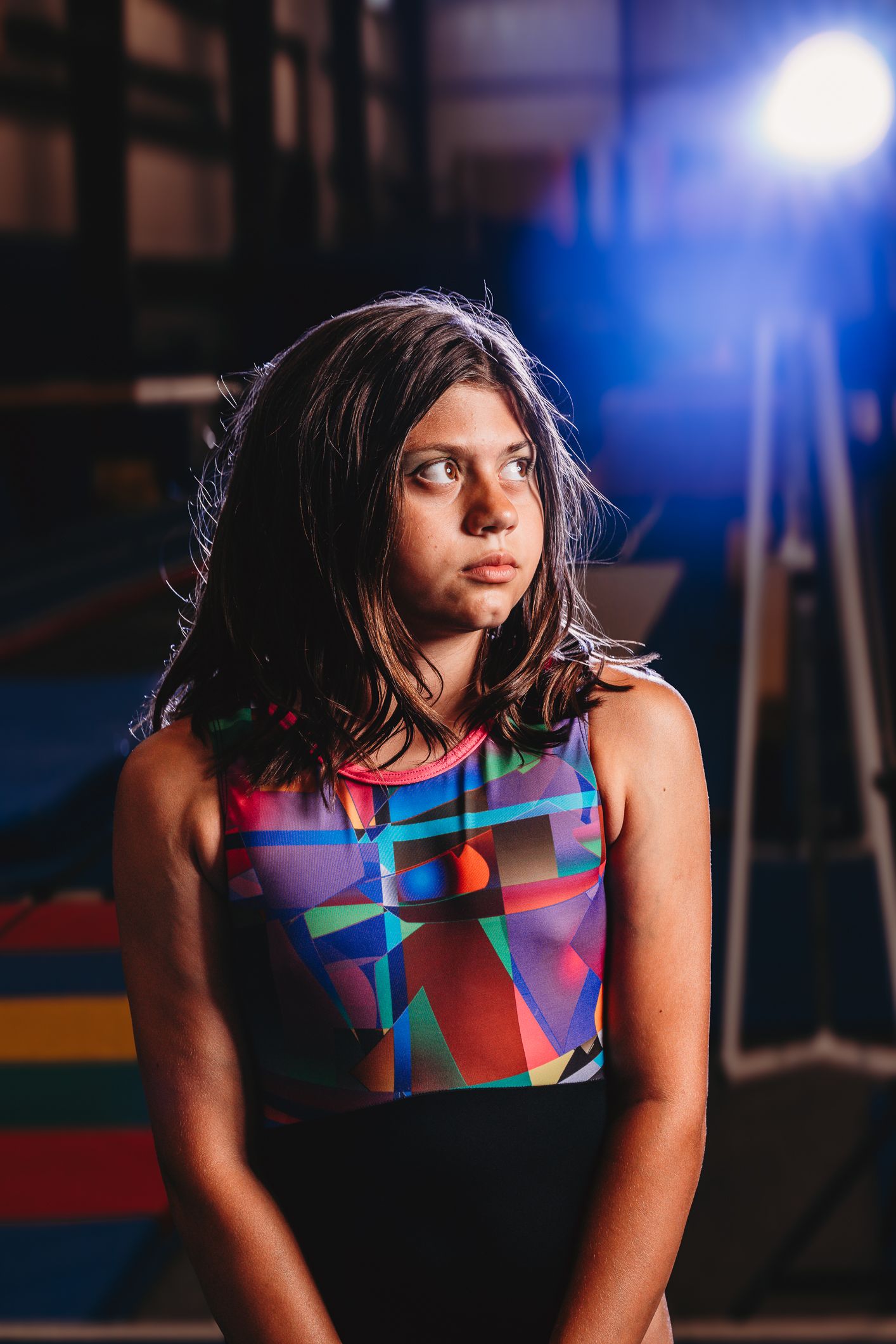 Young gymnast in colorful leotard stands in dimly lit gymnasium with dramatic blue light in background.