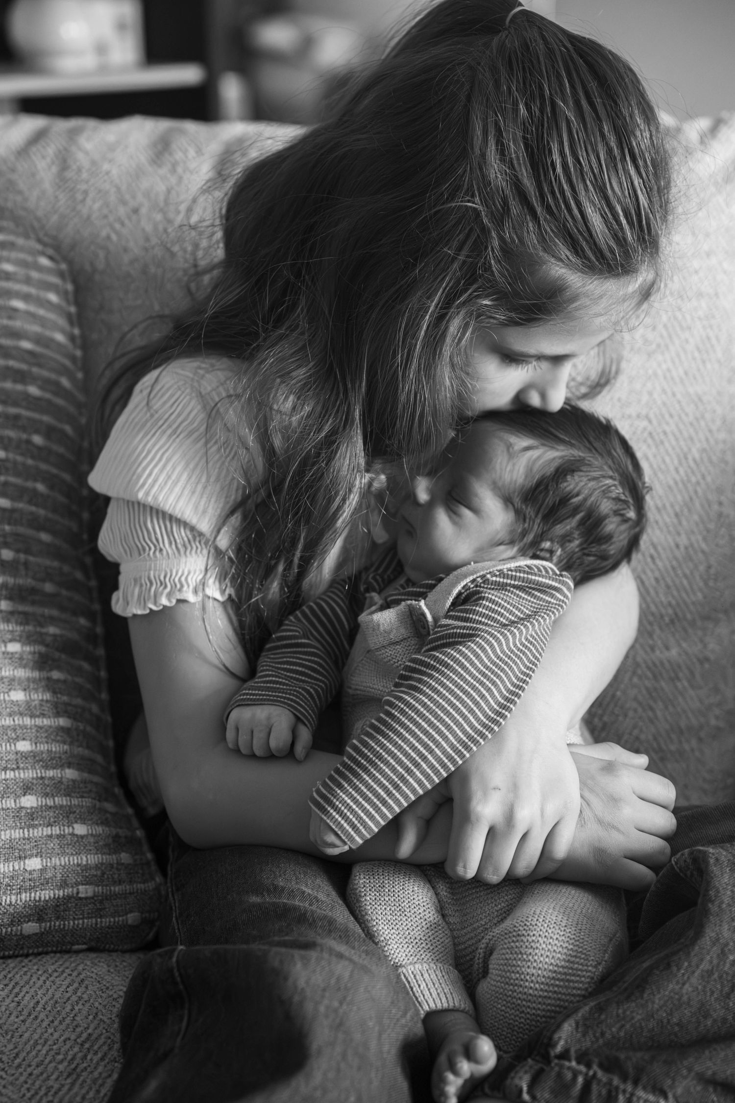 Black and white photo of a young girl tenderly holding and kissing a newborn baby on a cozy sofa.