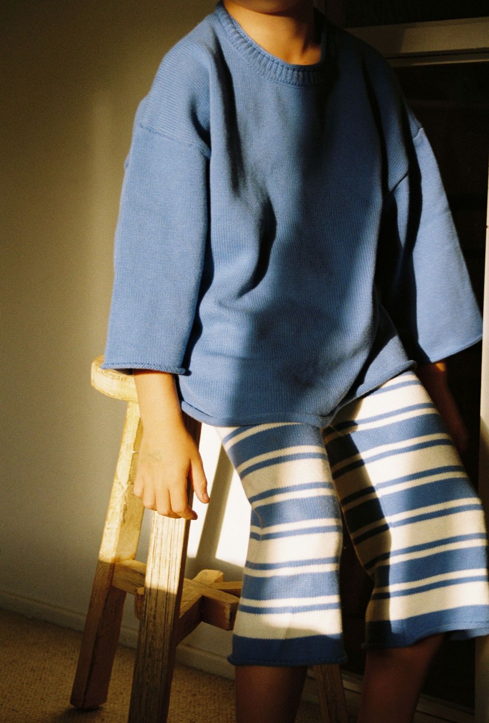 Child wearing a blue oversized knit sweater and striped blue and white skirt, standing beside a wooden stool.