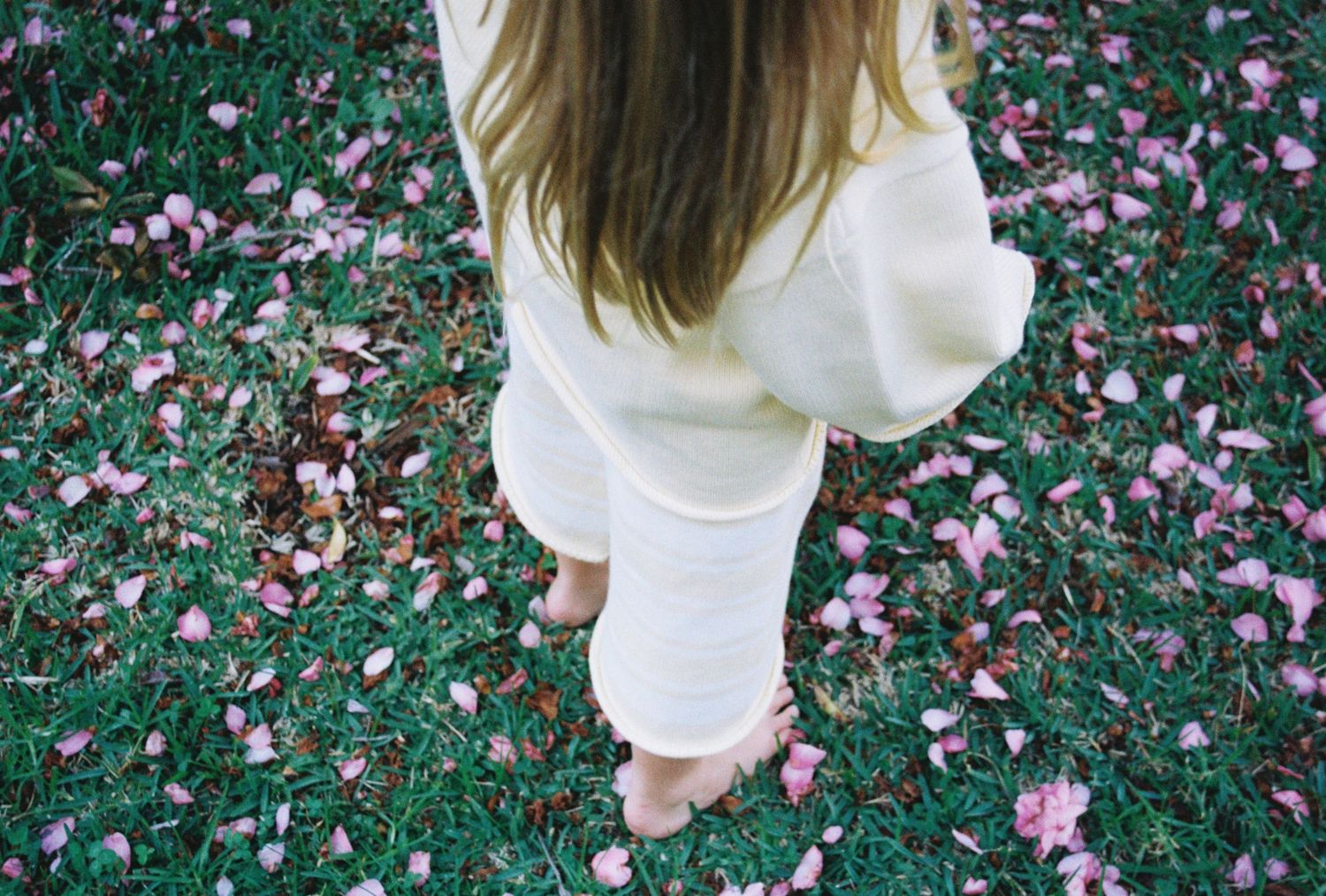 Person in white outfit standing barefoot on green grass scattered with pink flower petals.