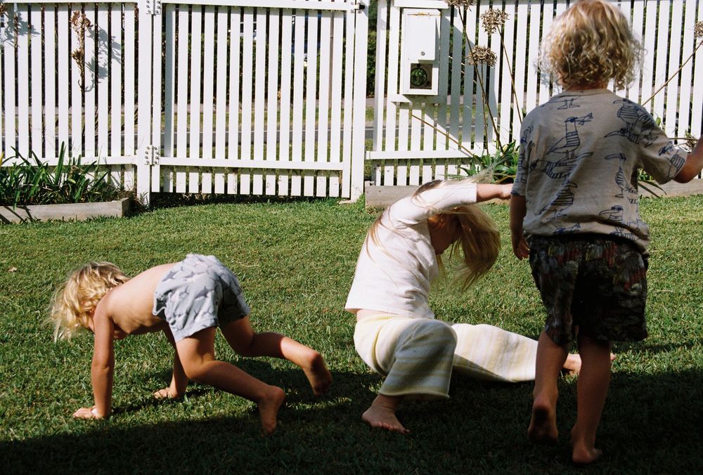 Three children playing barefoot on green grass in a backyard with a white picket fence.