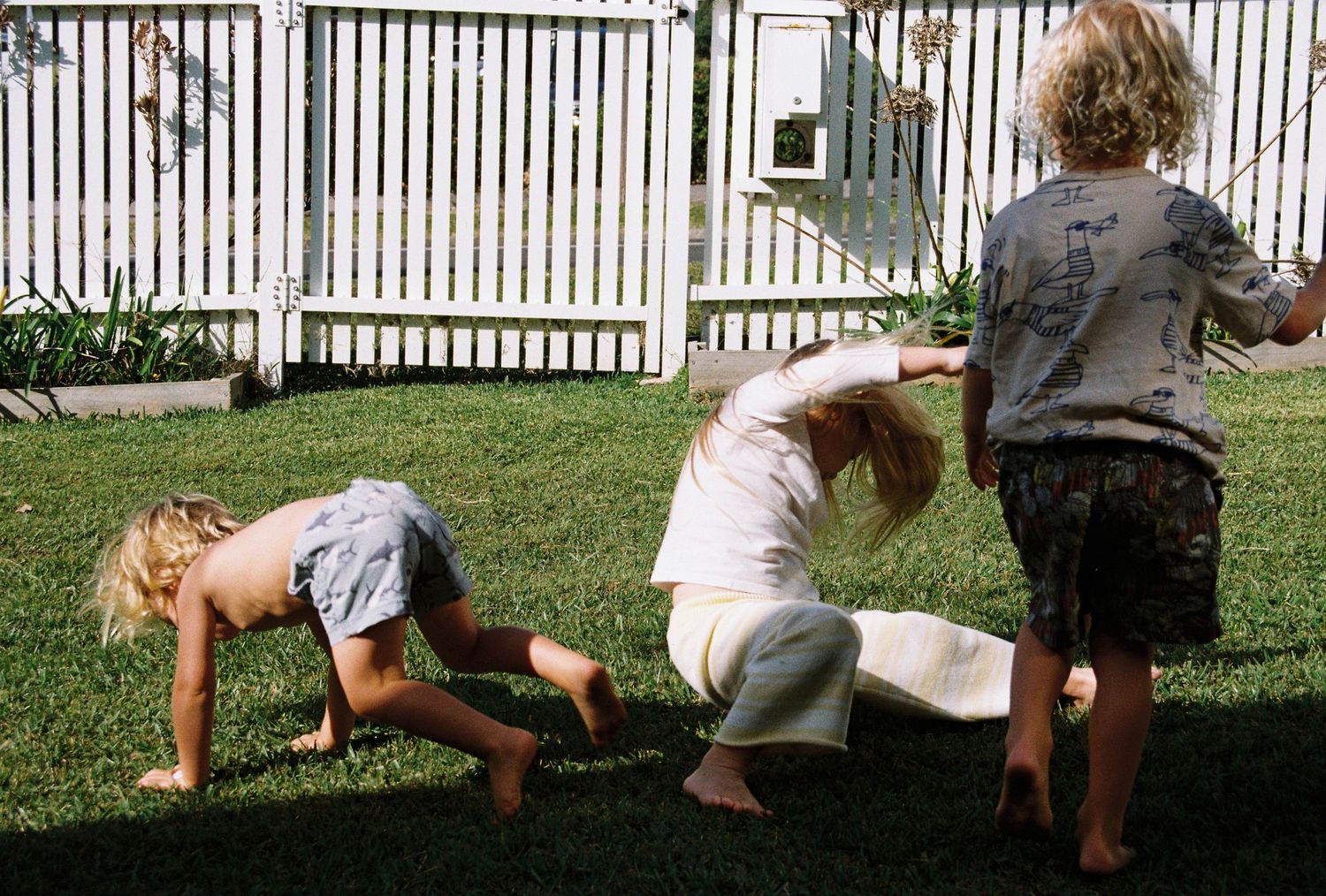 Three children playing barefoot on green grass in a backyard with a white picket fence.