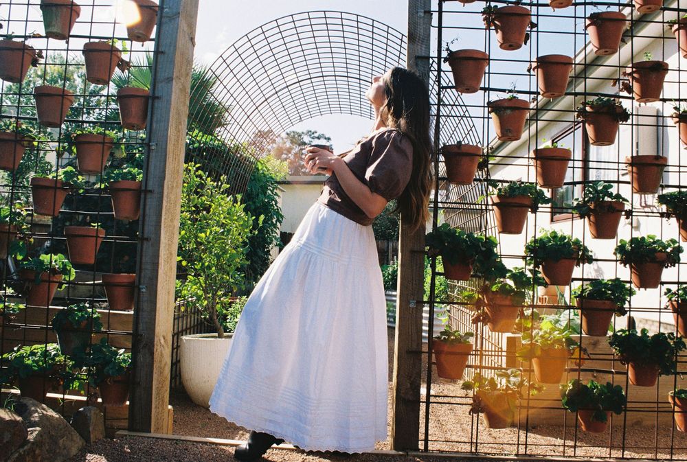 Woman in white skirt stands by a garden gate surrounded by terracotta pots on a wall trellis.