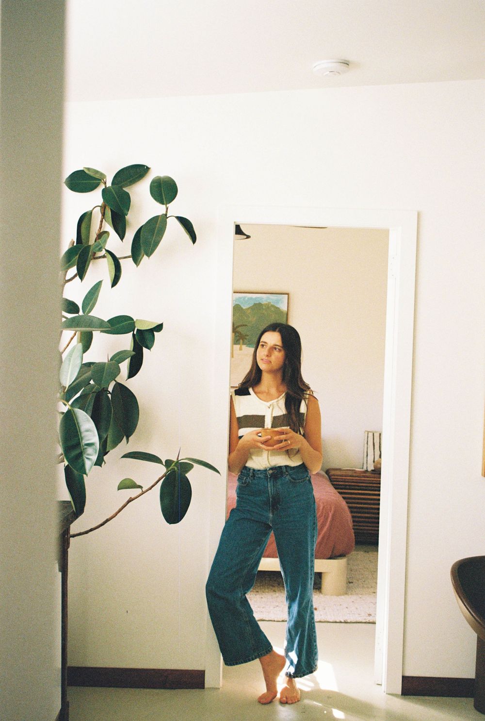 Woman in white top and blue jeans stands barefoot in doorway beside large green plant in bright modern home.