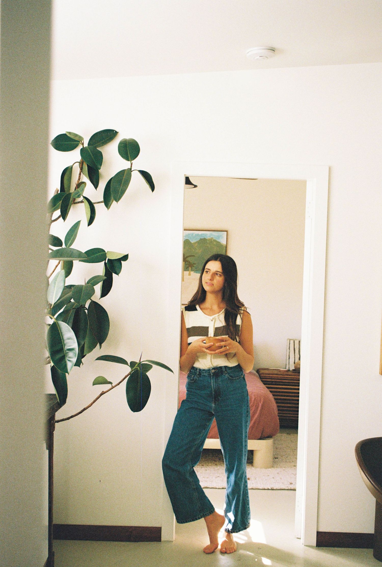 Woman in white top and blue jeans stands barefoot in doorway beside large green plant in bright modern home.