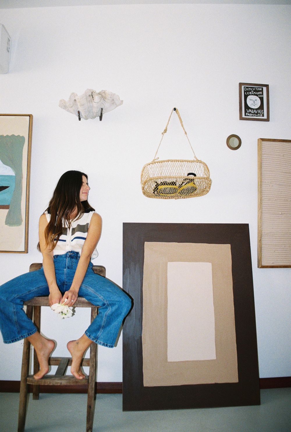 Woman in jeans sits on stool in art studio with paintings, wicker basket, and decorative wall pieces.