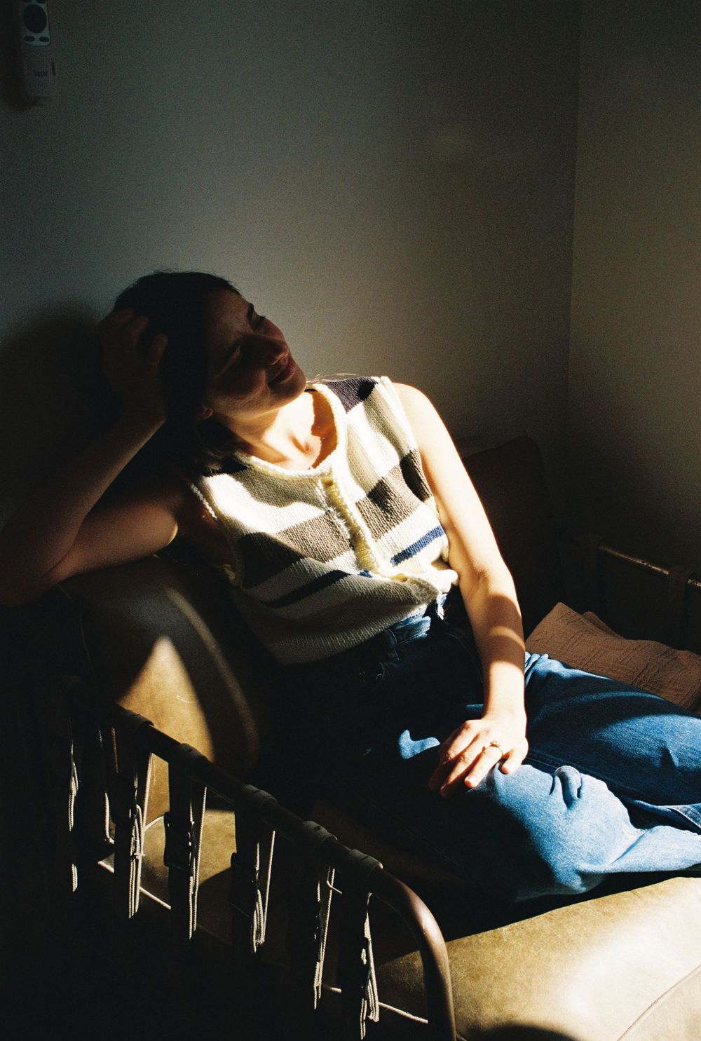 A person reclines in a chair bathed in dramatic sunlight, wearing a striped vest and jeans, hand raised to their head.