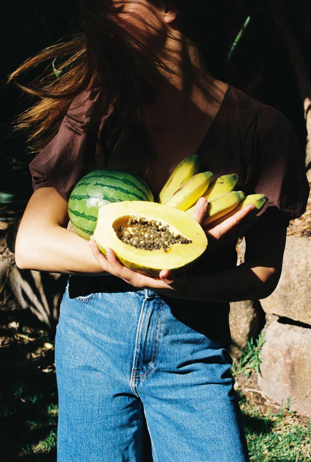 Woman in blue jeans holding a halved papaya, bananas, and a watermelon outdoors in sunlight.