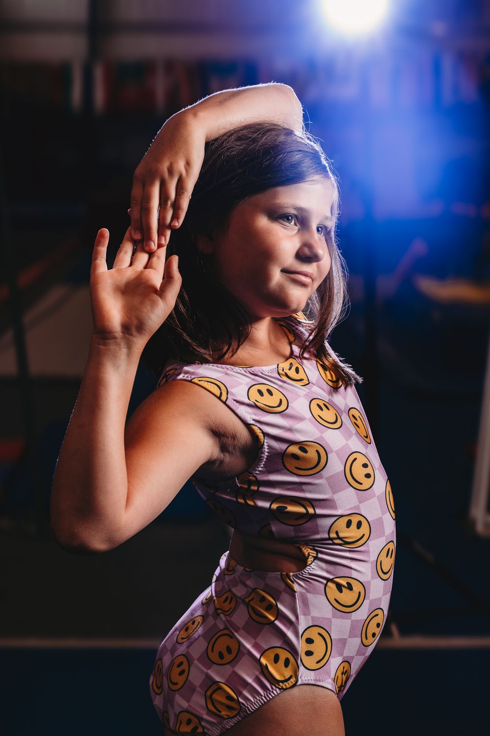 Young girl in smiley face leotard poses gracefully with raised hand in dimly lit gymnasium.