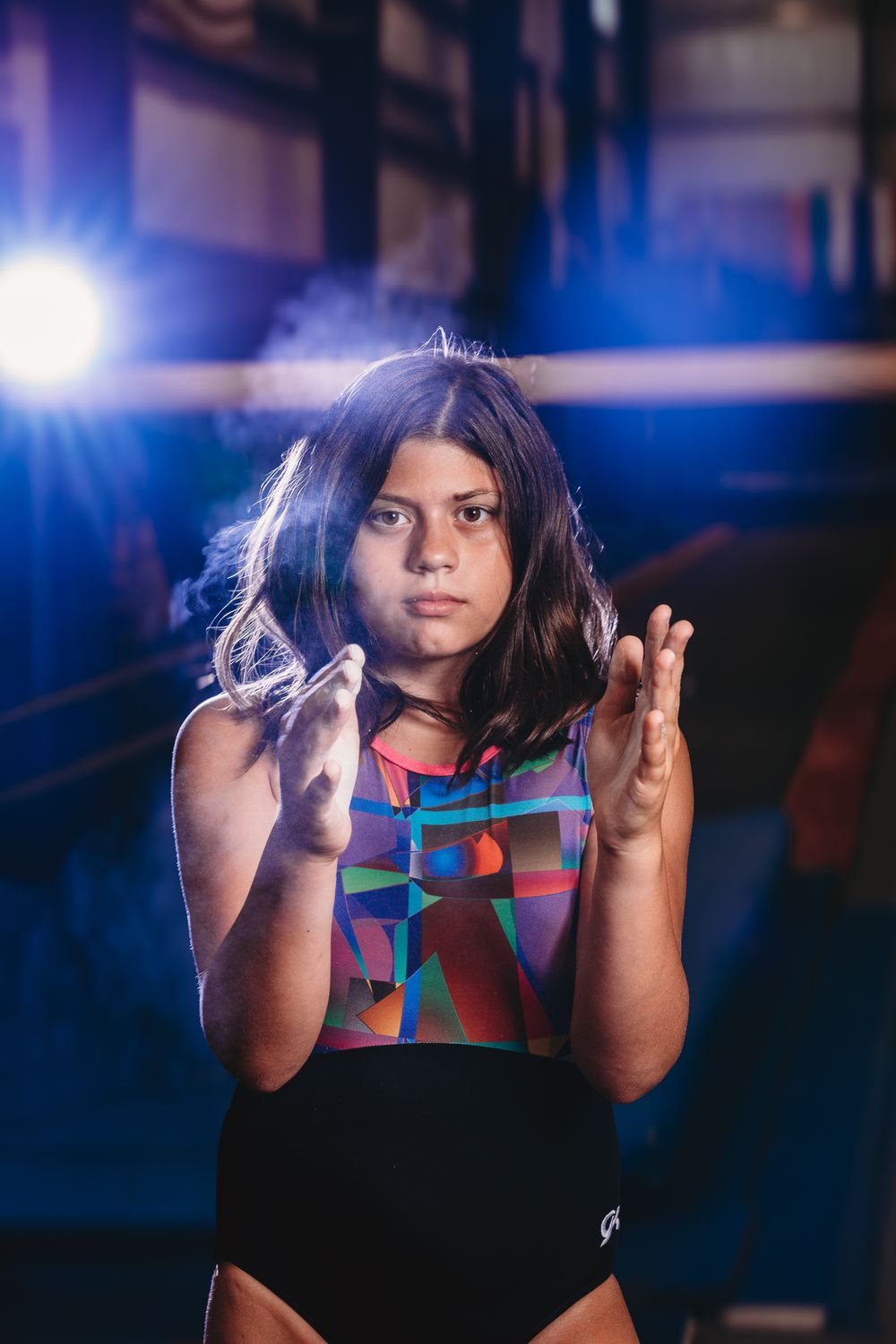 Young gymnast in colorful leotard clapping chalk from hands under blue stage lights in a gym.