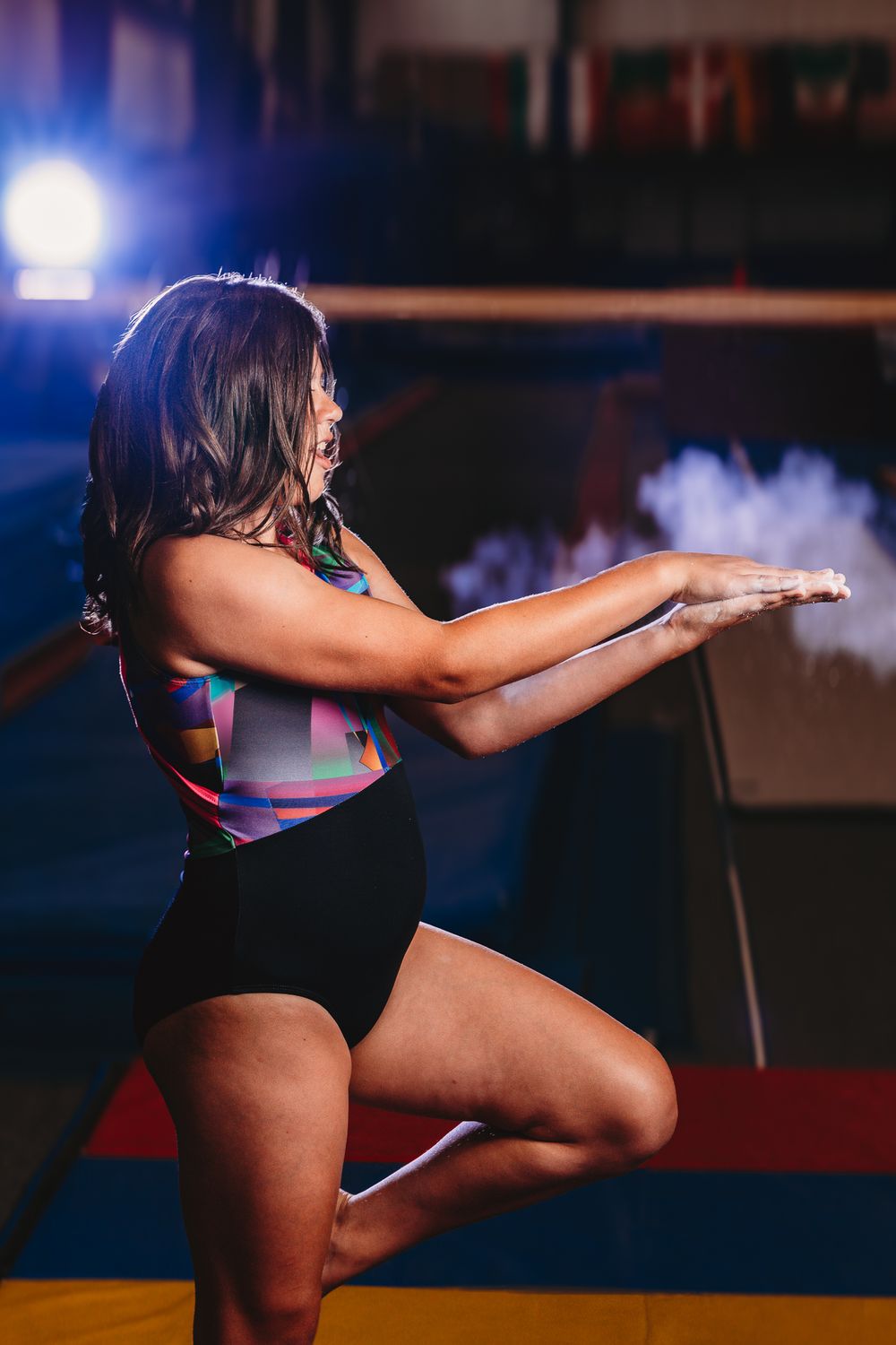 Female gymnast in colorful leotard balancing on one leg with arms extended, dusting chalk in a dimly lit gymnasium.