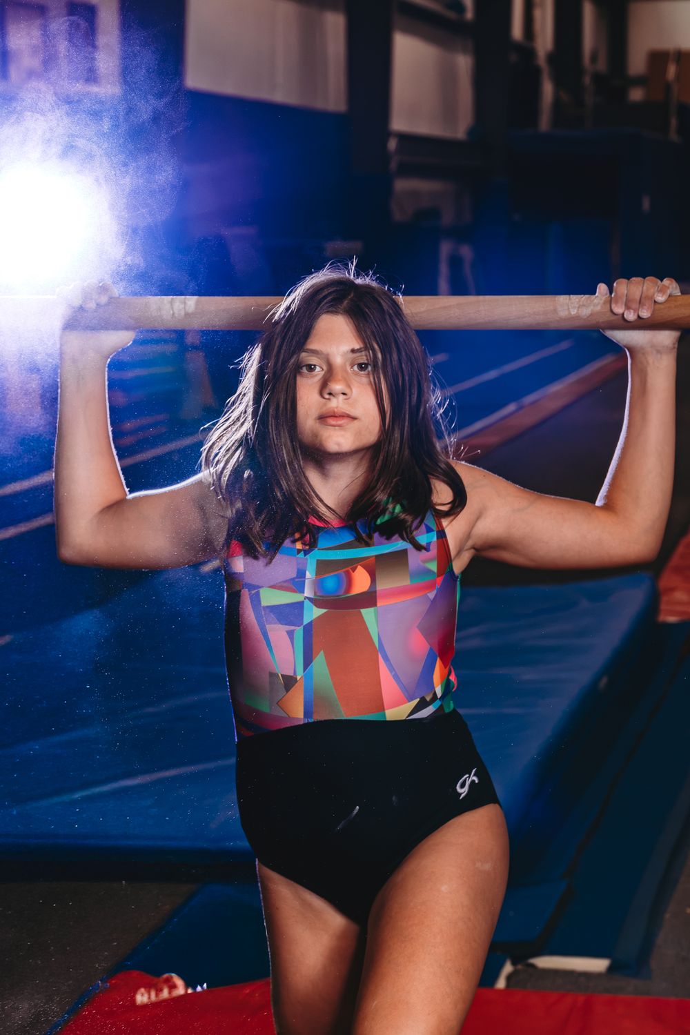 Young girl gymnast in multicolor GK leotard holds wooden bar across shoulders in dimly lit gymnastics training facility.