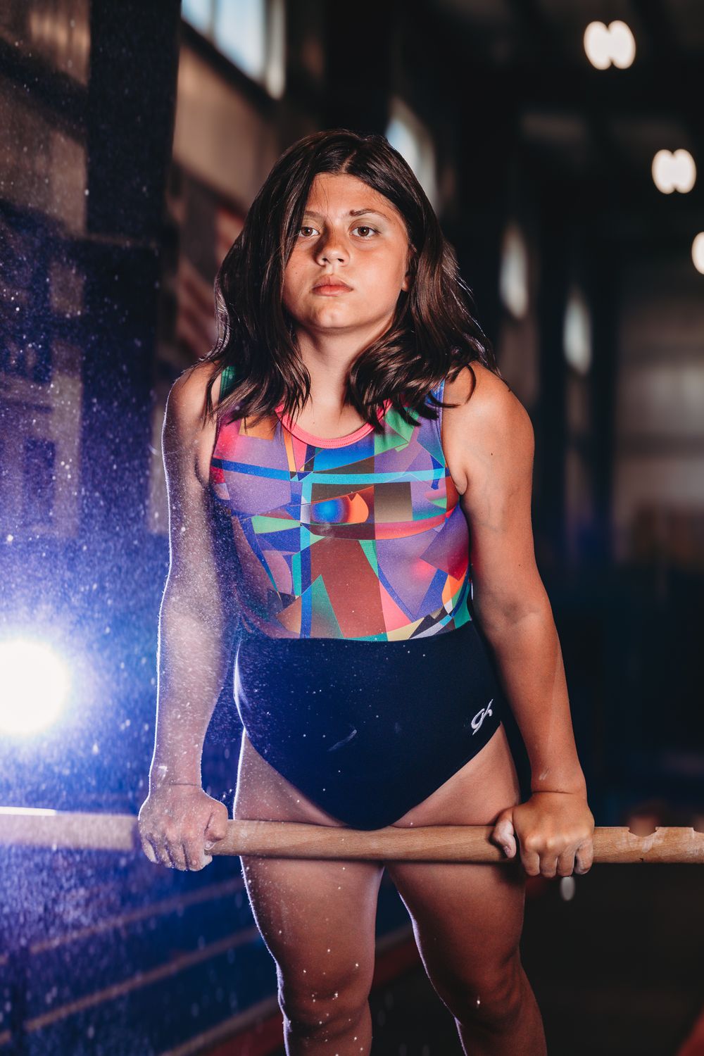 Young gymnast in colorful leotard gripping uneven bars with chalk dust in dimly lit gym.