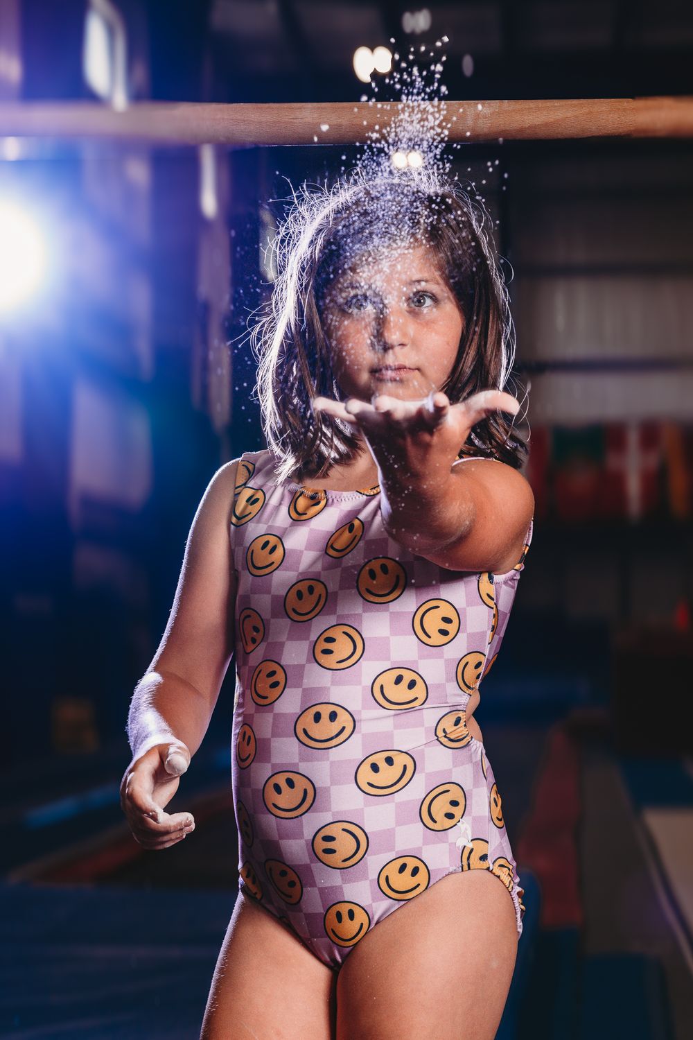 Young gymnast in smiley face leotard blows chalk dust from hands toward camera in dramatic lit gymnastics gym setting.