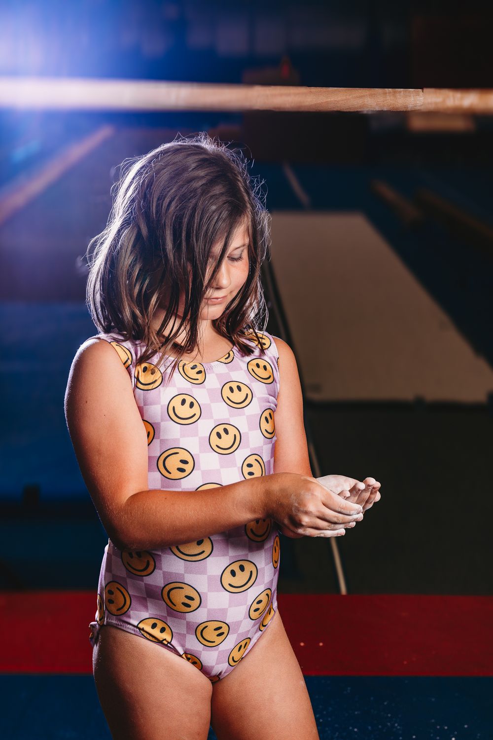 Young girl gymnast in smiley face leotard applying chalk to hands near balance beam in gymnasium.