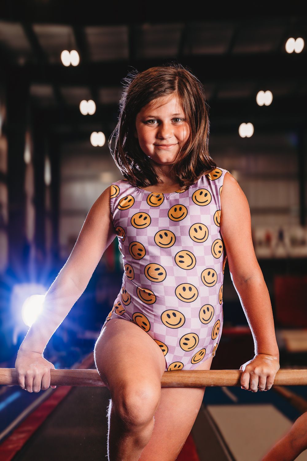 Young girl gymnast in smiley face leotard posing on parallel bars in a gym.
