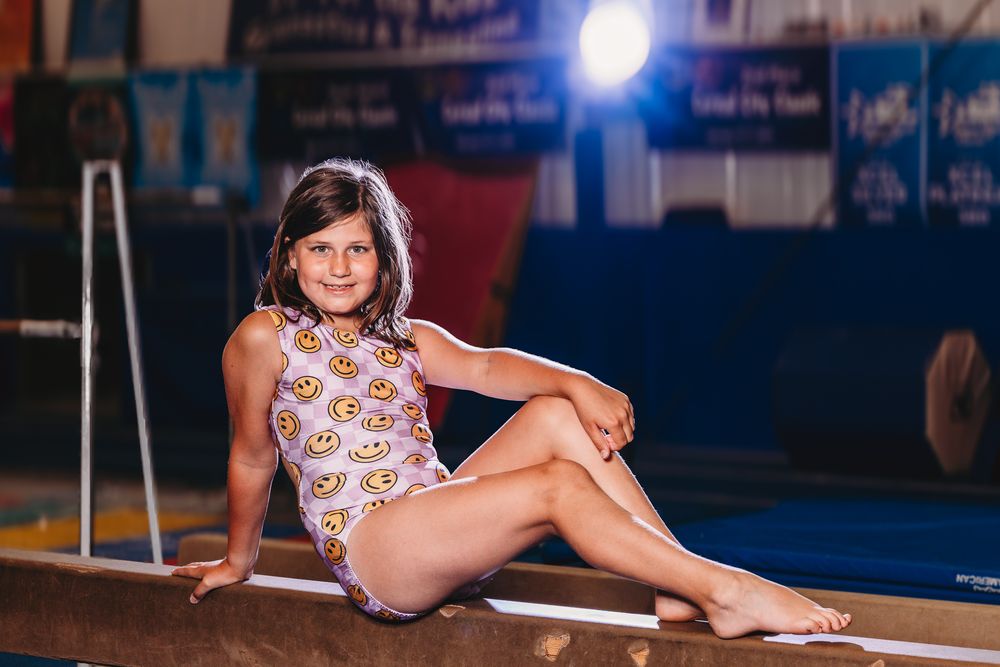 Young girl in patterned swimsuit sitting on gymnastics balance beam in indoor gym with bright lighting.